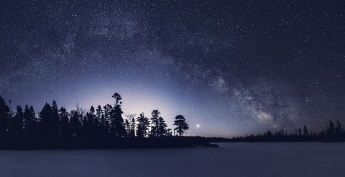 A starry night sky over a frozen lake, with silhouettes of trees and the Milky Way visible in the background.