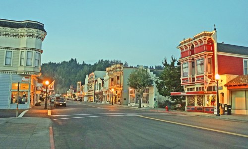 Charming small-town street at dusk, featuring historic buildings and warm streetlights amidst a forested backdrop.