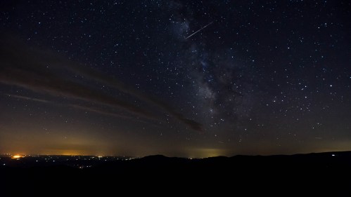 A starry night sky filled with the Milky Way and a shooting star over a dark landscape.