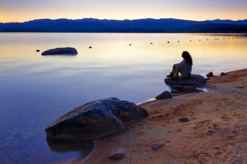 A person sits on a rock by a calm lake at sunset, with mountains in the background and a serene atmosphere.
