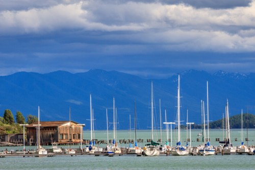 A serene harbor with sailboats docked, surrounded by mountains and a cloudy sky.