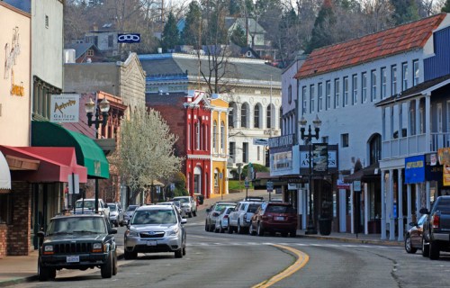 A charming street scene with shops, trees, and parked cars in a small town, featuring colorful buildings and a distant hill.