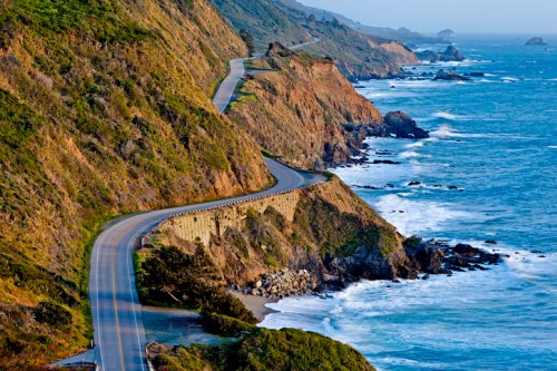 A winding coastal road hugs the cliffs above the ocean, with waves crashing against the rocky shore below.