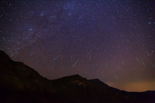 A starry night sky filled with shooting stars over dark mountain silhouettes.