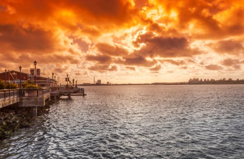 A vibrant sunset over a calm waterfront, with dramatic orange clouds and silhouettes of buildings along the shore.