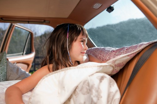 A young girl smiles while resting in a car, surrounded by pillows and blankets, looking out the window.