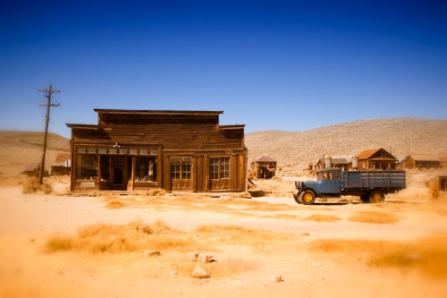 A rustic wooden building in a desert landscape, with a vintage blue truck parked nearby under a clear blue sky.