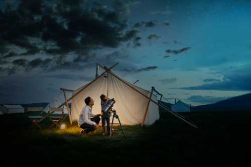 Two people observe the night sky through a telescope near a tent, surrounded by a starry sky and clouds.