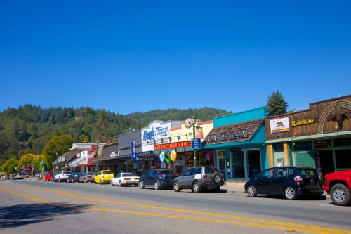 A sunny street lined with shops and parked cars, surrounded by green hills under a clear blue sky.
