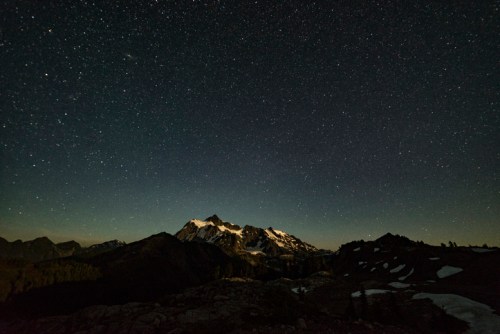 A starry night sky over a mountain range, with peaks illuminated by a soft glow and a clear view of the stars.