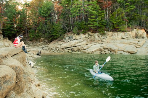 A person in a kayak paddles on a calm lake while another sits on a rocky shore, surrounded by trees.