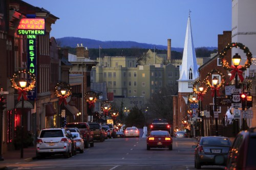 A festive street scene at dusk, with holiday decorations, cars, and a church steeple in the background.
