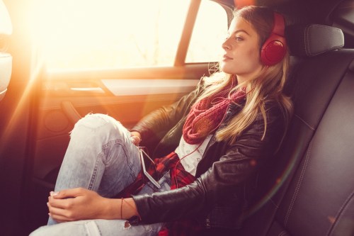 A young woman with headphones relaxes in the backseat of a car, enjoying the sunlight streaming through the window.