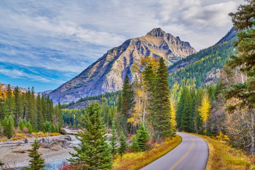 A scenic road winds through a forested landscape with mountains and a river, showcasing autumn colors.