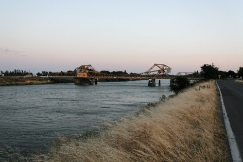 A wooden bridge spans a river at dusk, with dry grass lining the riverbank and a road nearby.