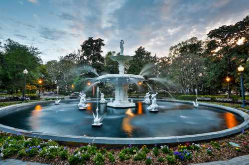 A white fountain with statues surrounded by flowers, set in a park under a cloudy sky.