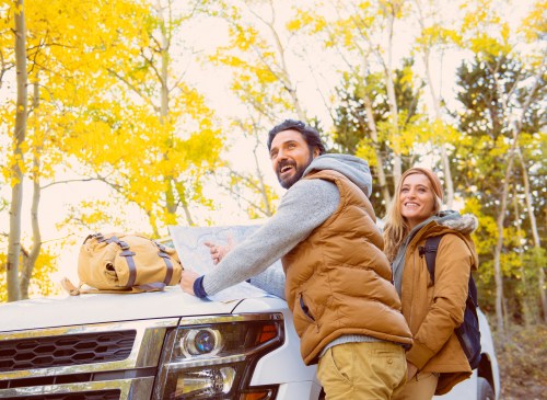 A couple smiles while looking at a map beside a white SUV, surrounded by vibrant autumn trees.