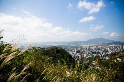 A panoramic view of a cityscape surrounded by greenery and mountains under a blue sky with scattered clouds.
