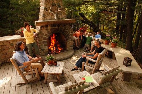 A group of people enjoying a cozy outdoor gathering around a stone fireplace in a wooded setting.