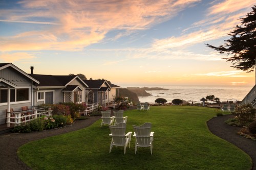Cozy cottages by the ocean at sunset, with a grassy area and white chairs overlooking the water.