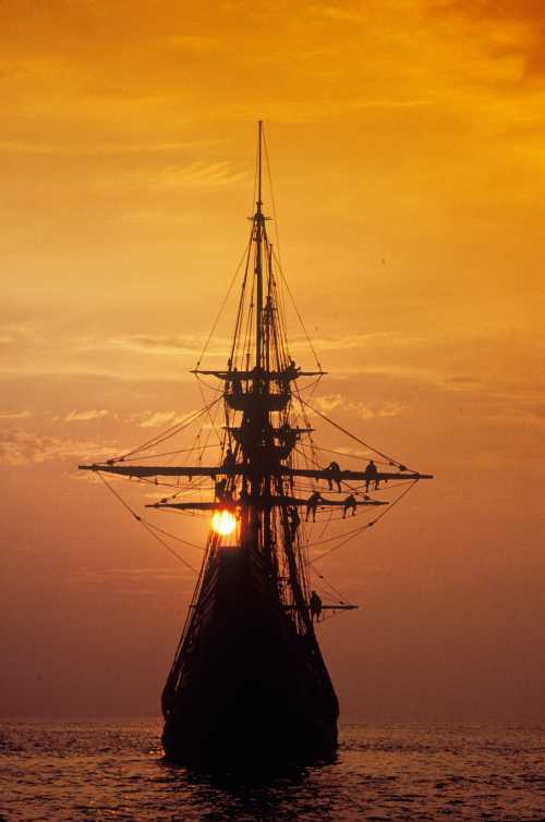 Silhouette of a tall ship against a vibrant orange sunset, with crew members visible on the rigging.