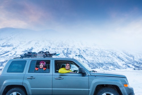Two people in a car with snow-covered mountains in the background, enjoying a winter adventure.