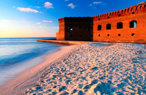 A sandy beach with calm waters and a historic brick structure at sunset, creating a serene coastal scene.