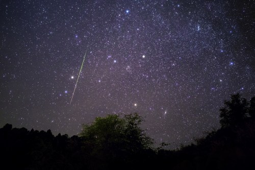 A starry night sky filled with stars and a bright meteor streaking across the scene above a dark landscape.
