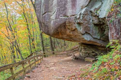 A large rock formation overhangs a dirt path, surrounded by colorful autumn trees and a wooden railing.