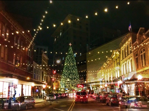 A festive street scene at night, featuring a large Christmas tree and twinkling lights strung above the bustling road.