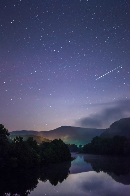 A serene night sky filled with stars above a calm river, reflecting mountains and a shooting star in the distance.