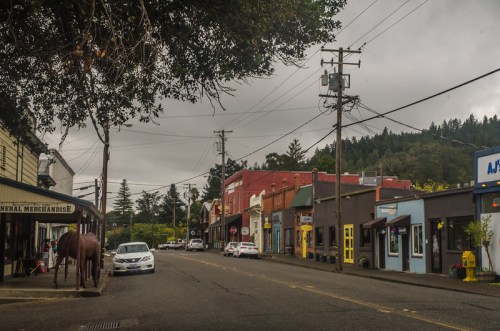 A quiet street scene featuring small shops, power lines, and a horse statue, set against a cloudy sky and green hills.