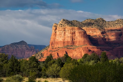 Red rock formations rise majestically against a blue sky, surrounded by green trees and distant mountains.