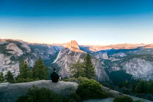 A person sits on a rock, overlooking a vast mountain landscape at sunset, with trees in the foreground.