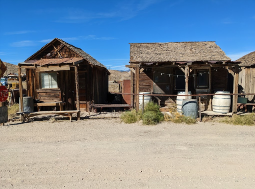 Two rustic wooden buildings with a dirt path, barrels, and a clear blue sky in the background.
