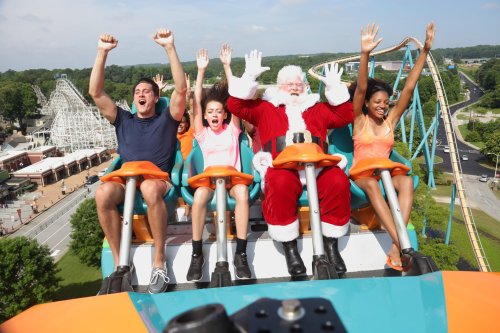 A group of four people, including Santa, joyfully ride a roller coaster with hands raised in excitement.