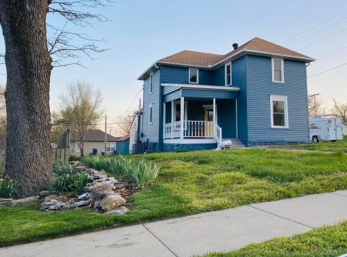 A blue two-story house with a porch, surrounded by green grass and a small garden, under a clear sky.
