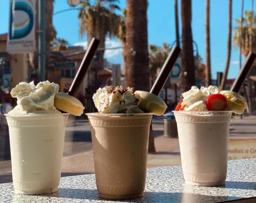 Three milkshakes in cups topped with whipped cream and fruit, set against a sunny palm tree backdrop.