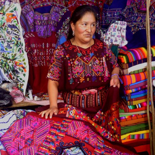 A woman in traditional attire stands among colorful textiles and woven fabrics in a market setting.