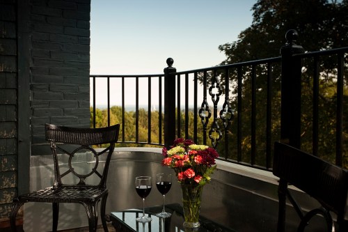 A balcony with two chairs, a glass table, a vase of flowers, and two glasses of red wine, overlooking a scenic view.
