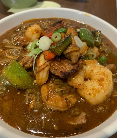 A bowl of spicy seafood stew with shrimp, vegetables, and herbs, served in a rich brown broth.