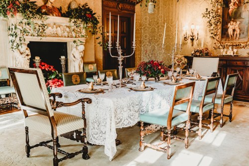 Elegant dining table set with lace tablecloth, candles, and floral arrangements in a beautifully decorated room.