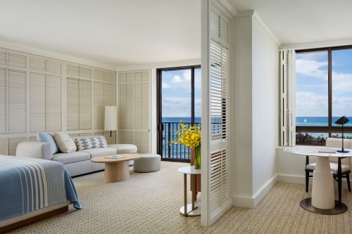 Bright hotel room with a view of the ocean, featuring a sofa, table, and large windows with natural light.