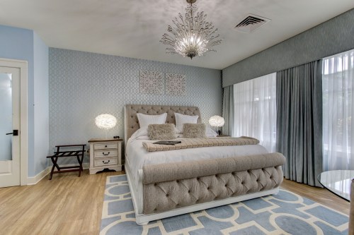A stylish bedroom featuring a tufted bed, elegant lighting, and soft blue decor with large windows and a patterned rug.