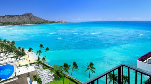 A stunning view of a turquoise ocean, palm trees, and a mountain under a clear blue sky.
