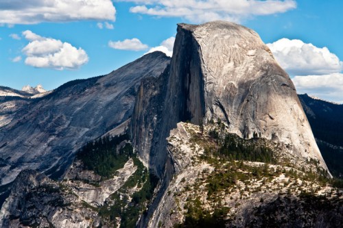 A majestic granite mountain peak under a blue sky with fluffy clouds, surrounded by rugged terrain and greenery.