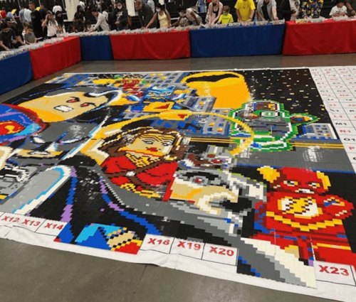 A large pixel art mural featuring superheroes, created with colorful blocks, displayed at a crowded event.