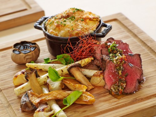A gourmet dish featuring sliced beef, roasted vegetables, and a small pot of creamy gratin on a wooden serving board.