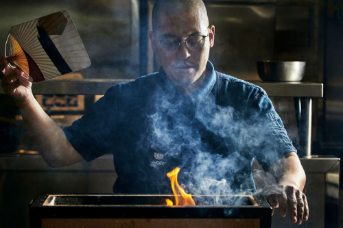 A chef in a blue uniform fans flames from a grill, surrounded by smoke in a kitchen setting.