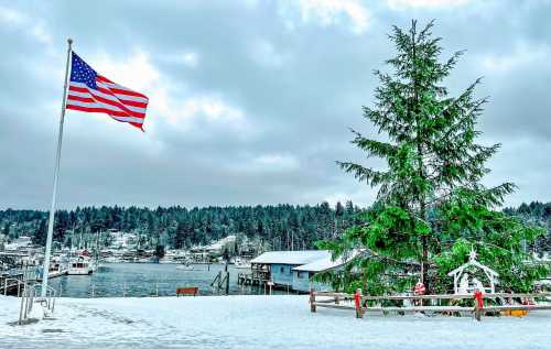 A snowy landscape featuring a Christmas tree, American flag, and a waterfront view with boats and pine-covered hills.